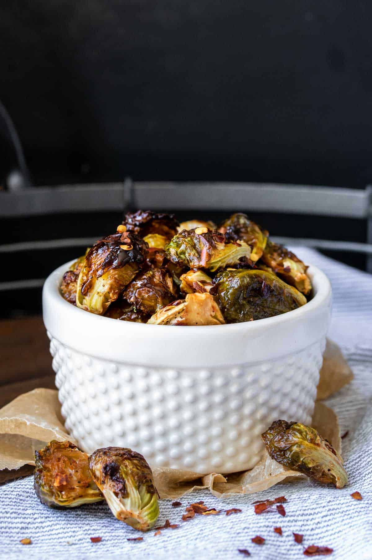 Brussels sprouts that have been roasted and browned in a white textured bowl sitting on parchment paper and a striped towel.