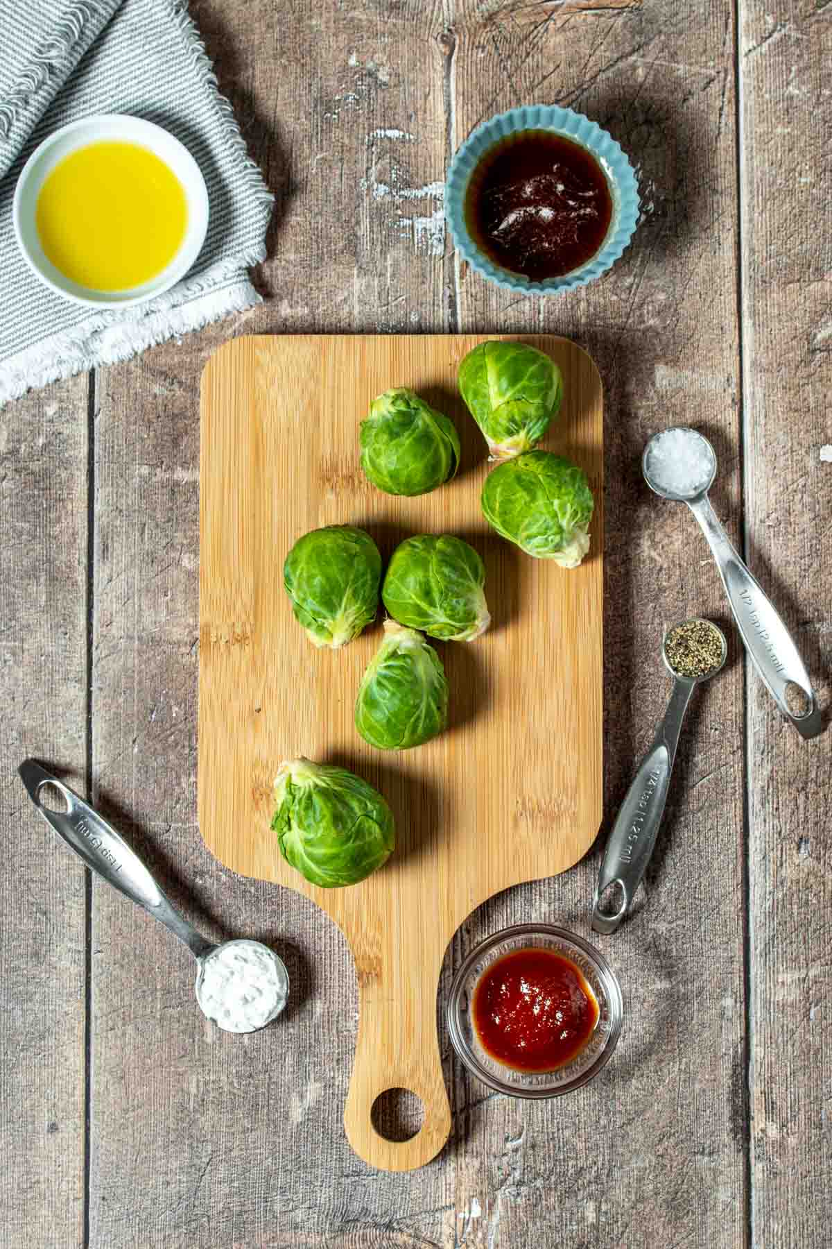 Top view of a wooden surface with Brussels sprouts on a wooden board surrounded by oil, hot sauce, maple syrup and seasonings.