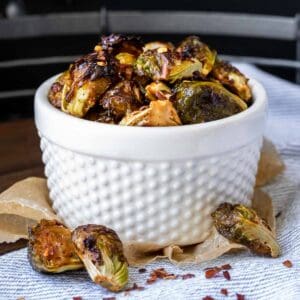 A white bowl with bumps on it filled with some browned roasted Brussels Sprouts and some fallen off in front.