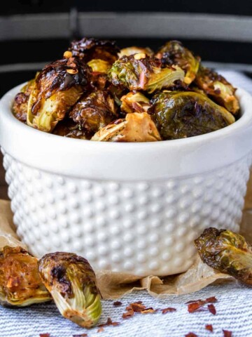 A white bowl with bumps on it filled with some browned roasted Brussels Sprouts and some fallen off in front.