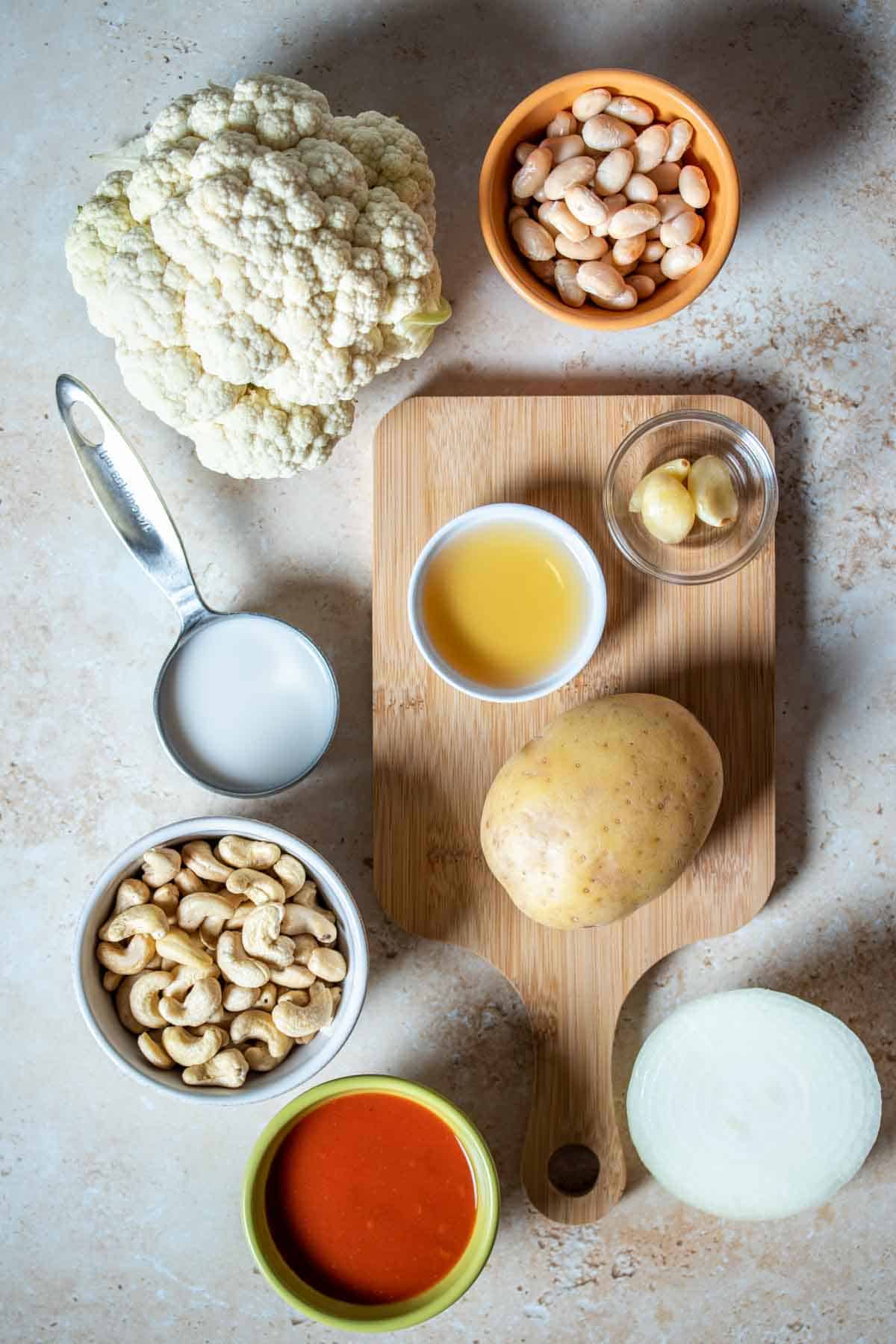 Top view of a tan surface with potatoes, cauliflower, onion, hot sauce, broth, milk, beans, nuts and garlic on it.