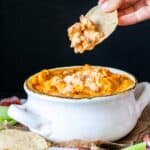 A hand with a tortilla chip dipping into a white bowl with buffalo chicken dip in it.