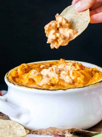 A hand with a tortilla chip dipping into a white bowl with buffalo chicken dip in it.