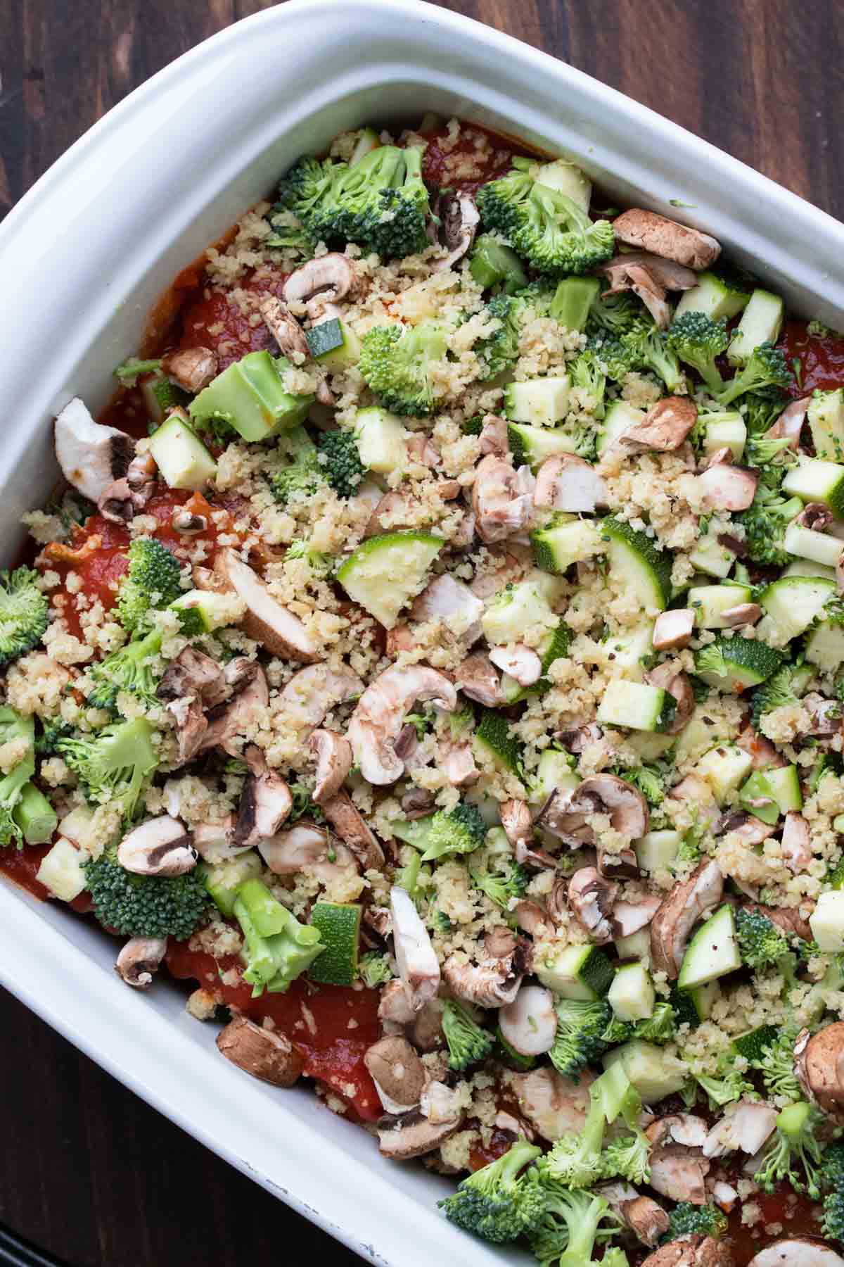 Chopped vegetables over red sauce and hidden lasagna layers in a white baking dish.