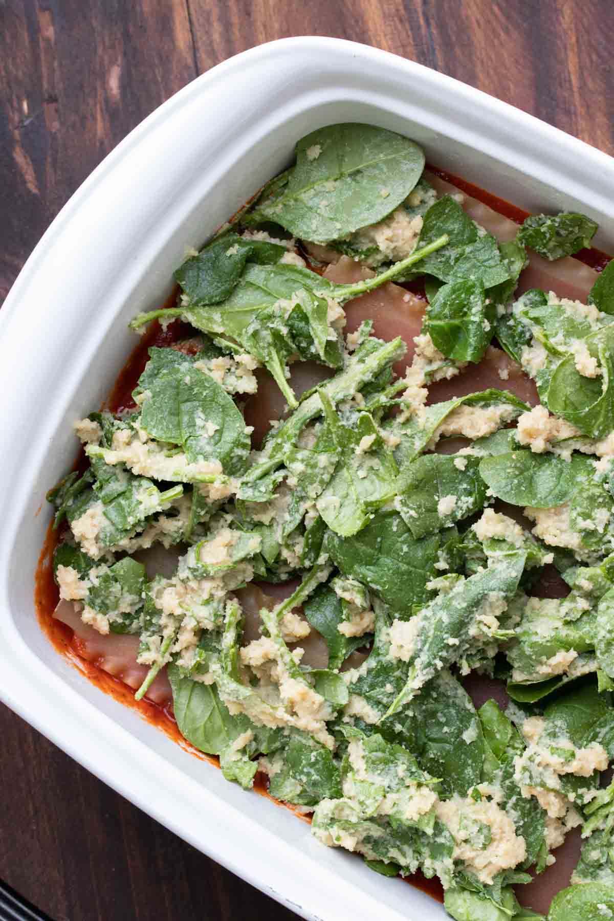 Top view of raw spinach over hidden layers of lasagna in a white baking dish.