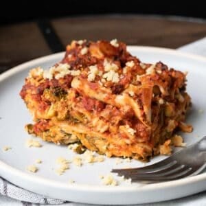 Front view of a piece of lasagna on a white plate with a fork on the plate.