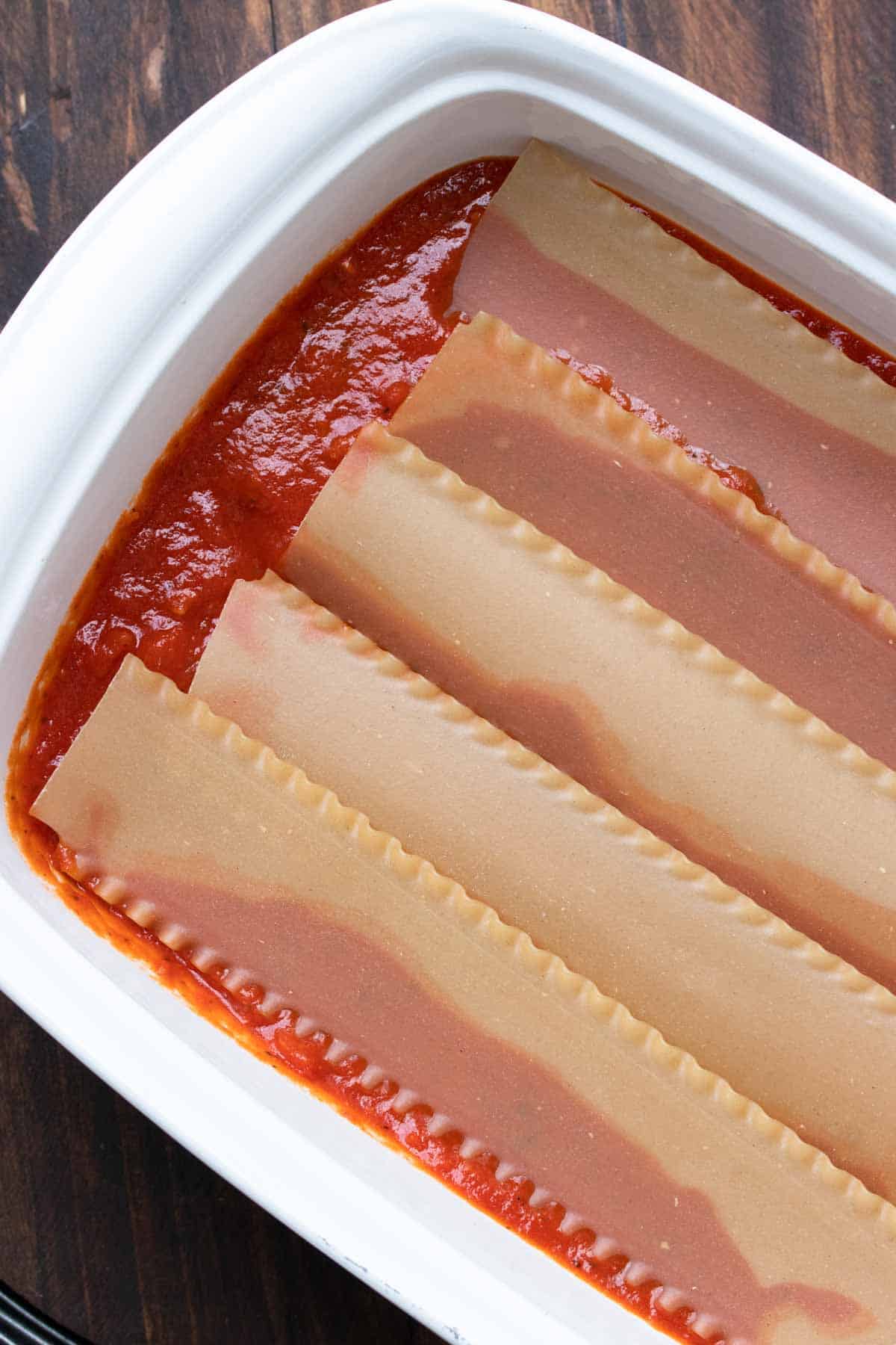 Top view of raw lasagna noodles in red sauce in a white baking dish.