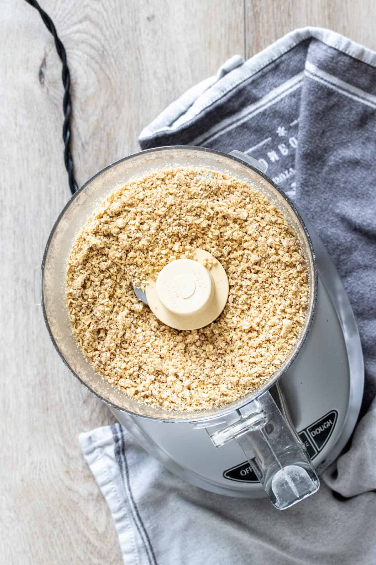 Top view of a food processor with chopped oats inside witting on a grey towel on a wooden surface.