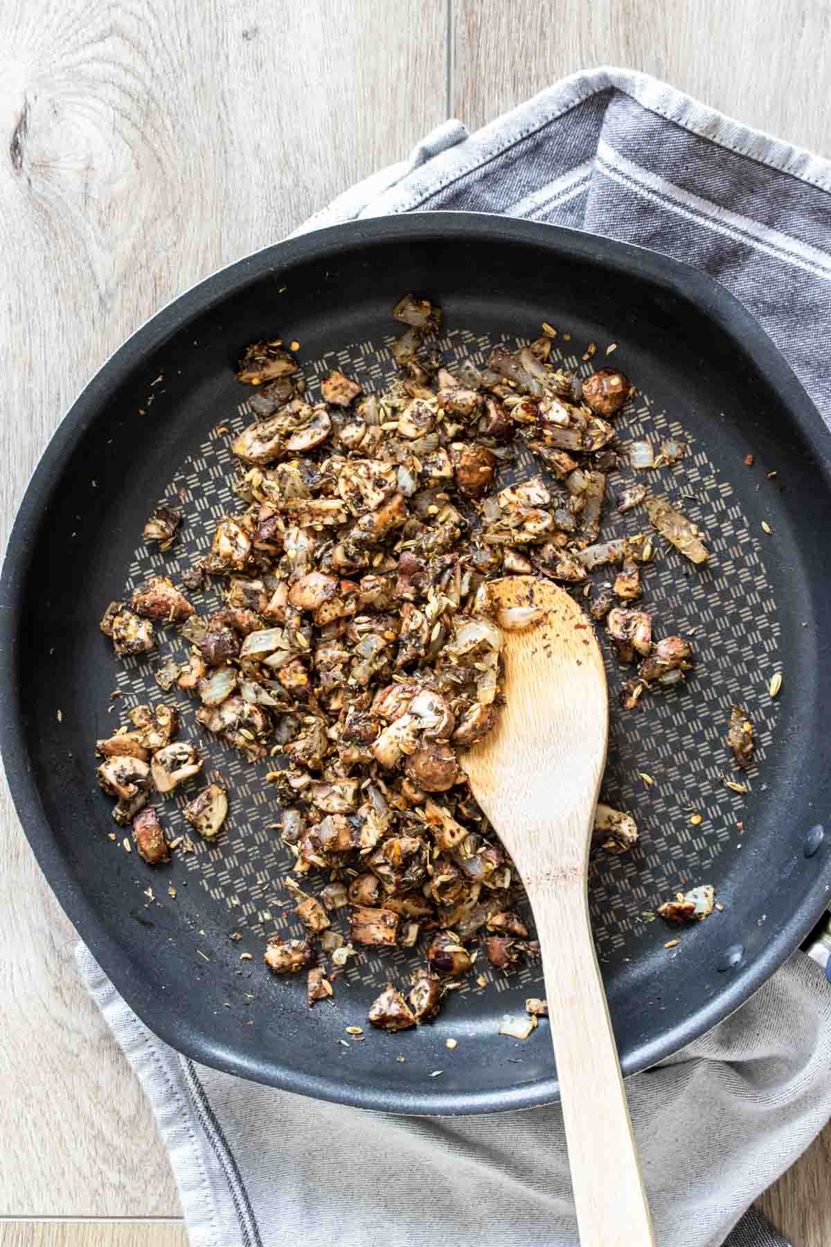 Chopped onions and mushrooms being sauteed in a black pan with a wooden spoon.