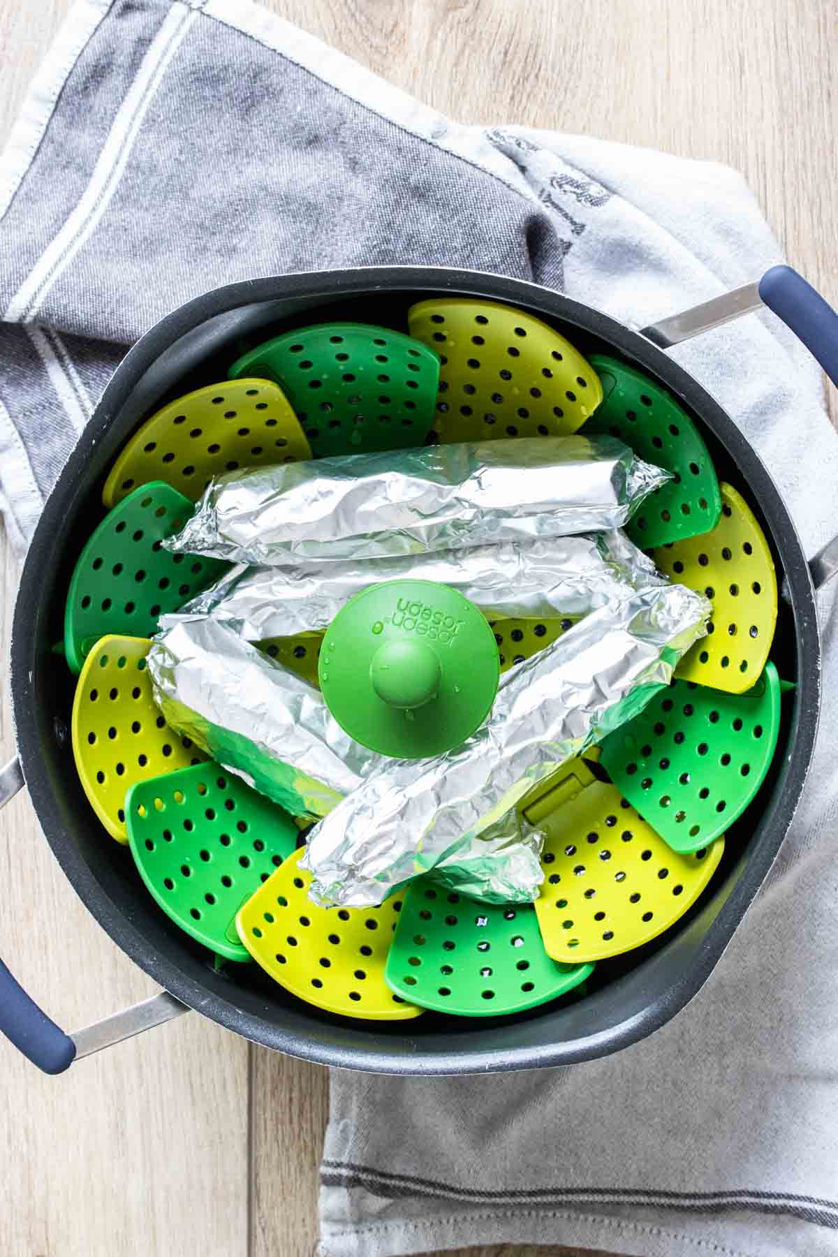 Top view of a green and yellow steamer basket in a black pot with four foil wrapped sausages in it.