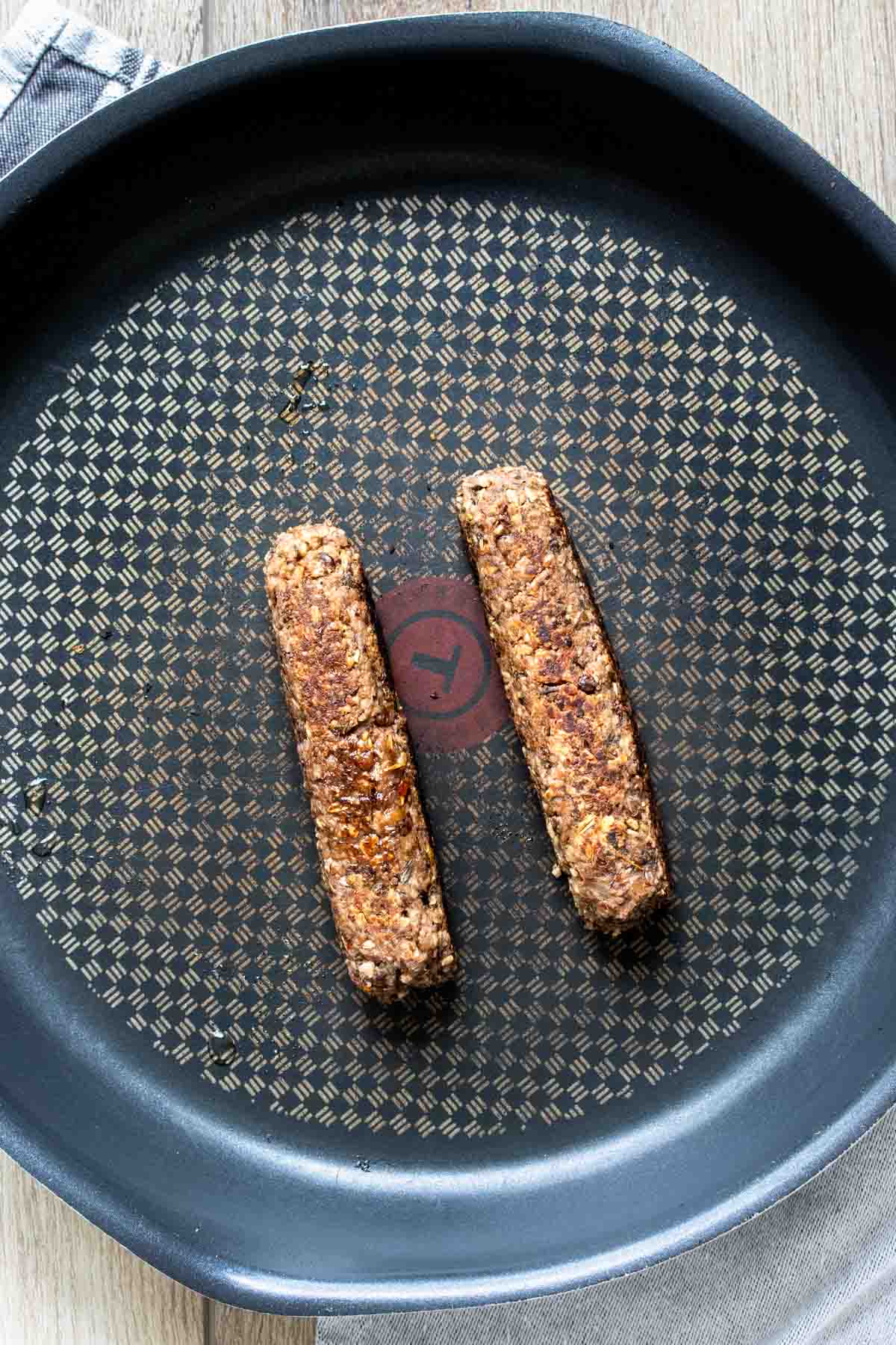 Two sausages in a black frying pan that is sitting on a wooden surface.