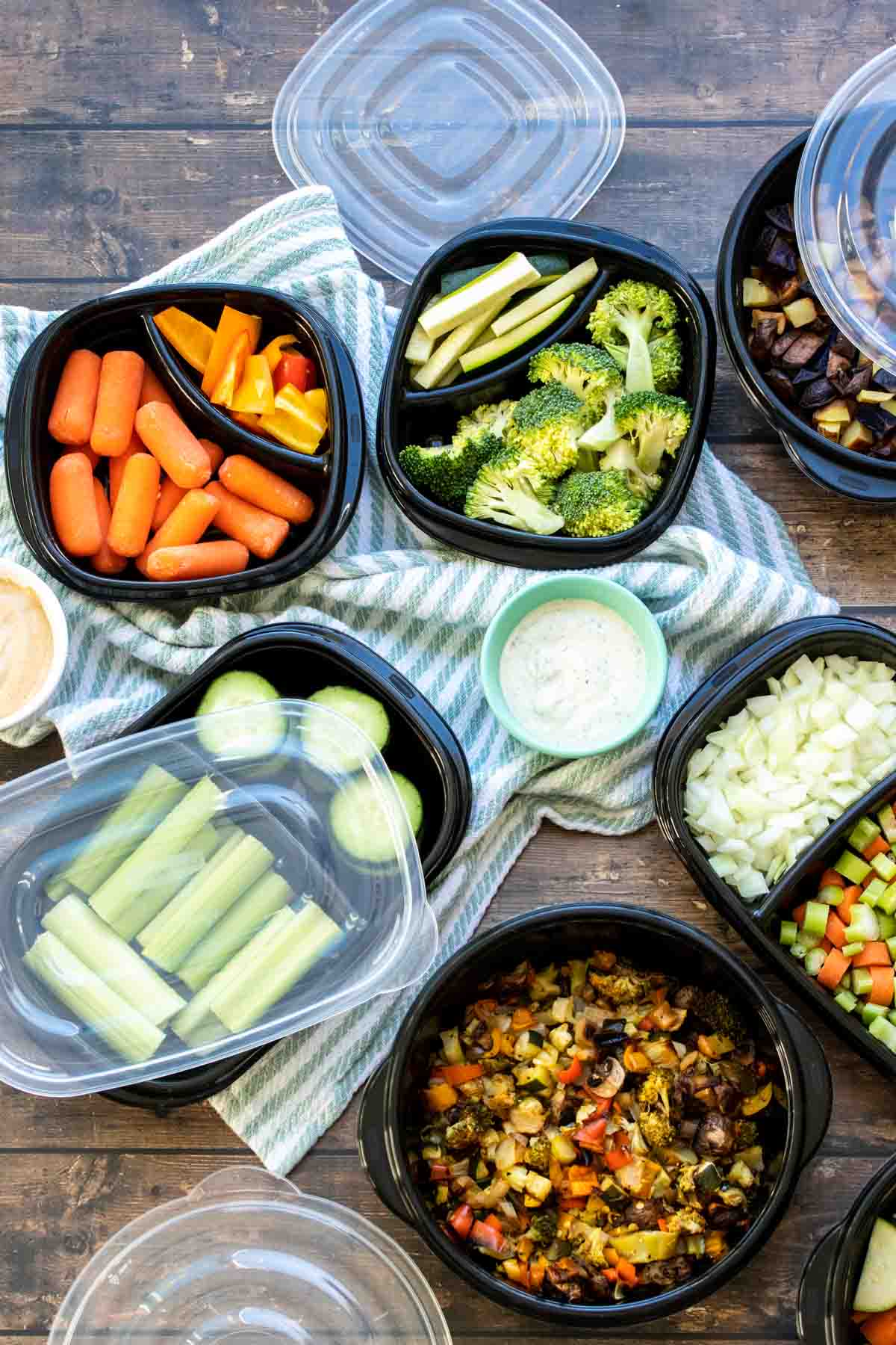 A wooden surface with a blue striped towel and black containers filled with fresh and roasted veggies in them.