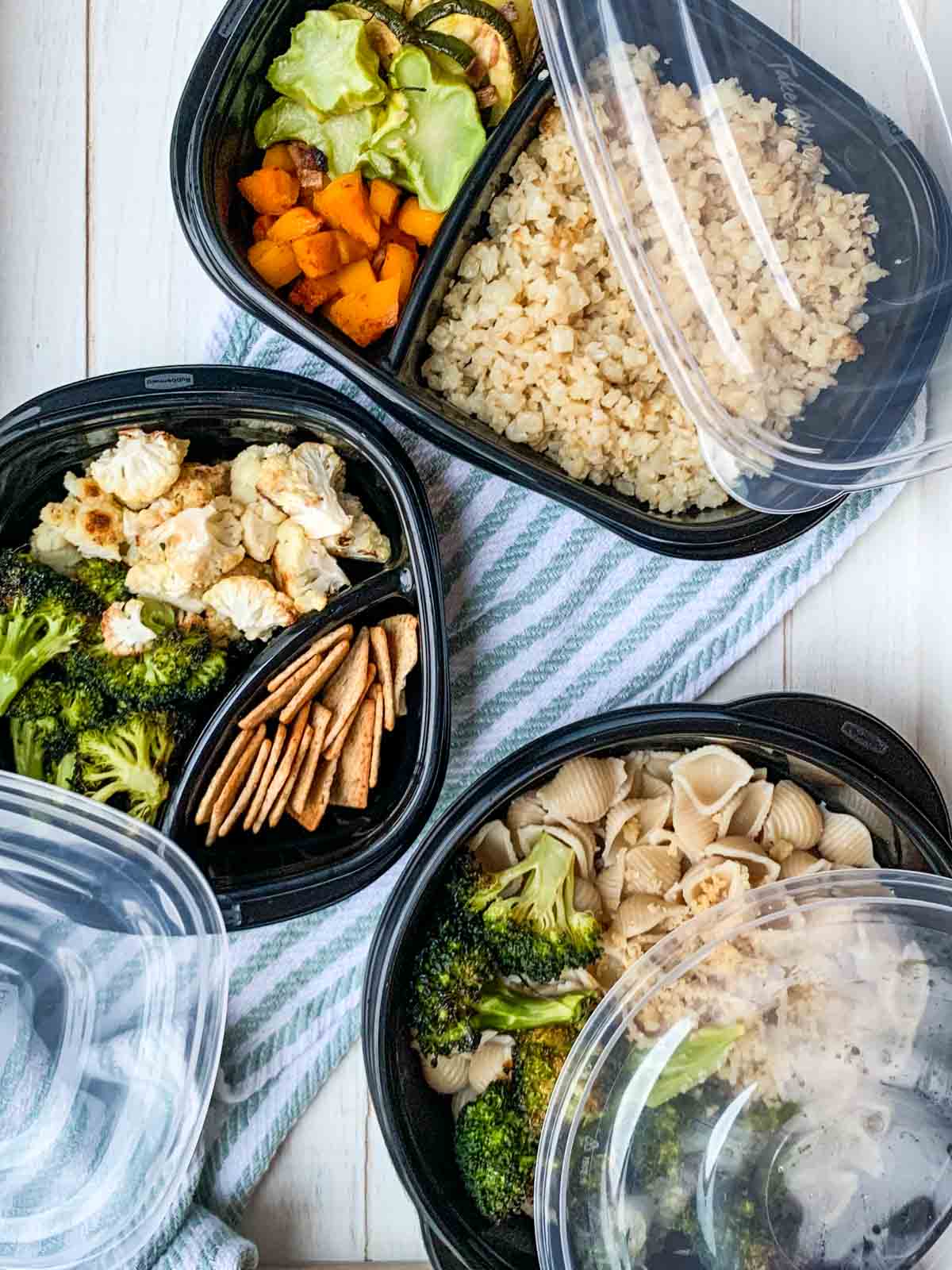 Three black containers with cooked veggies and different grains to make meals in them.