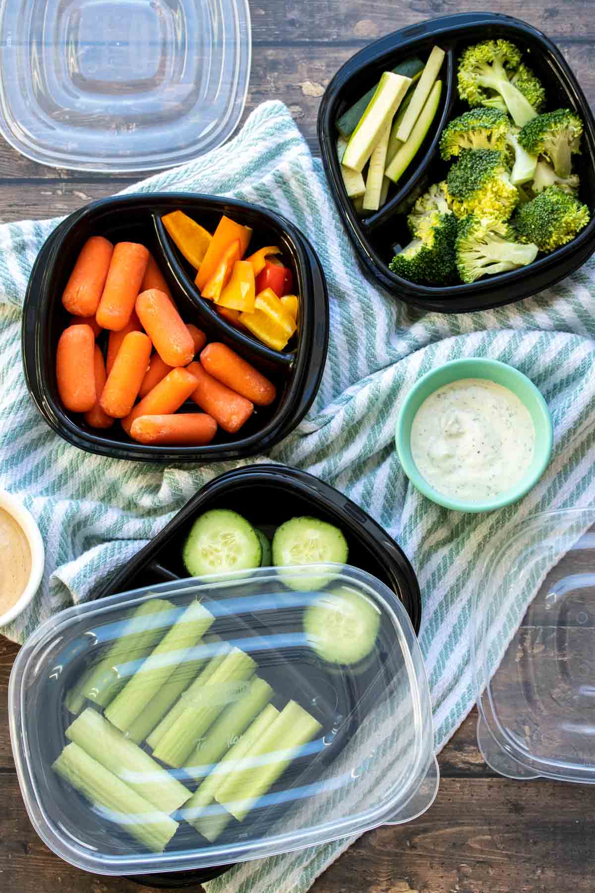 Fresh veggies in sectioned off black containers next to bowls of dips on a striped towel.