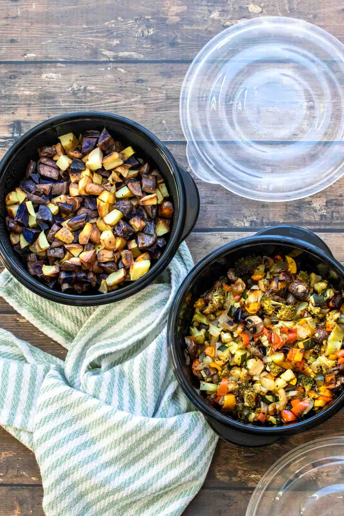 Top view of two black round containers filled with chopped roasted veggies sitting on a blue striped towel on a wooden surface.