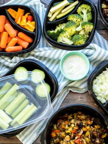 Fresh and cooked veggies in black containers of different sizes sitting on a striped towel with dips next to them.