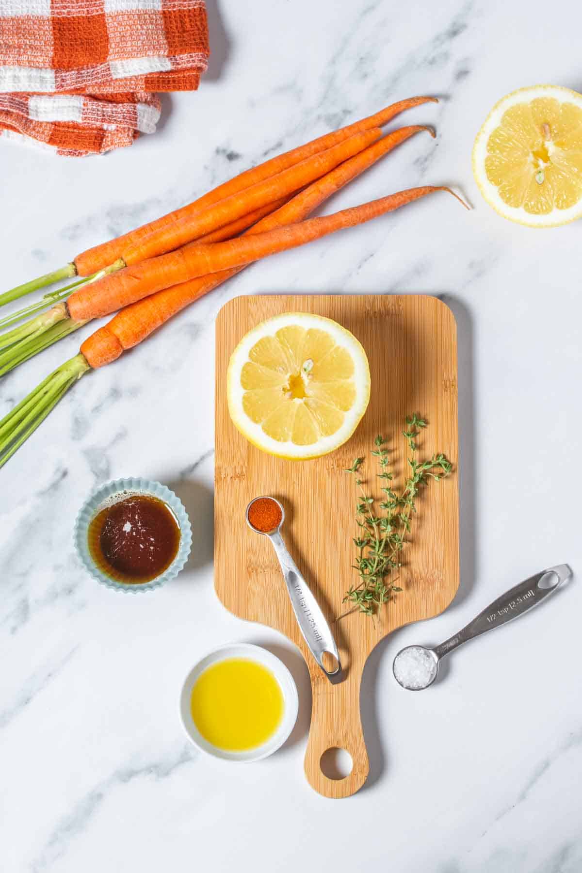 A wooden cutting board on a marble surface and thyme, seasonings, maple syrup, carrots, lemon and olive oil in bowls on them.