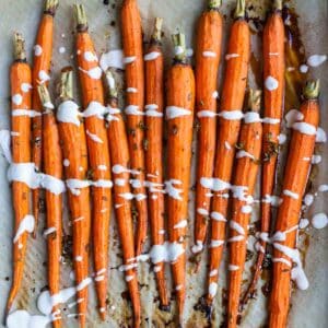 Roasted carrots lined up on parchment paper with brown spots on it and a white drizzle over the carrots.