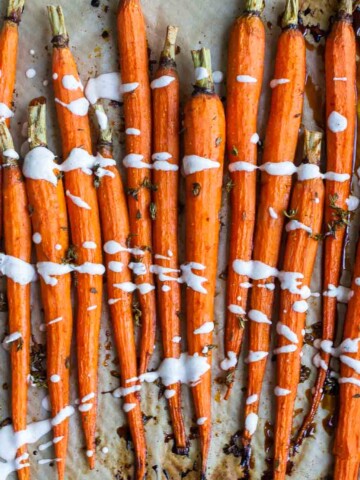Roasted carrots lined up on parchment paper with brown spots on it and a white drizzle over the carrots.