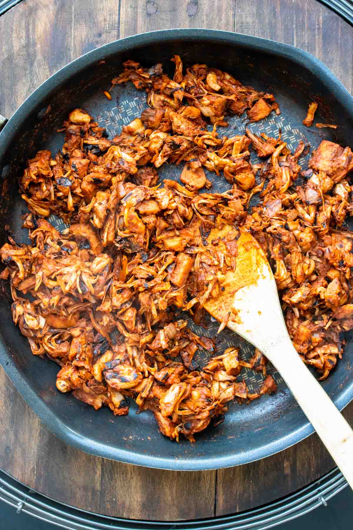 A wooden spoon mixing jackfruit mixed with red sauce in a black frying pan.