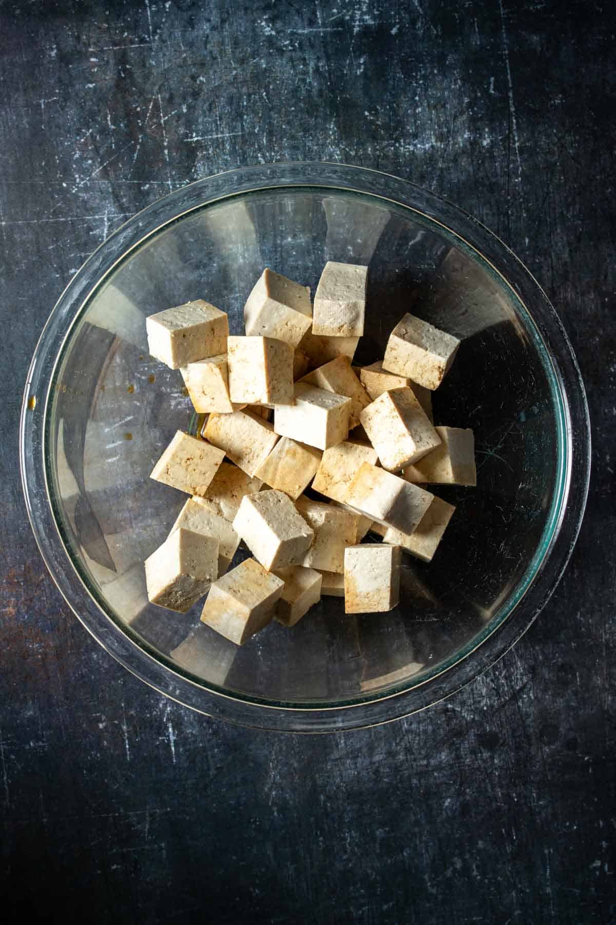 Top view of a glass bowl with pieces of tofu inside on a dark surface.