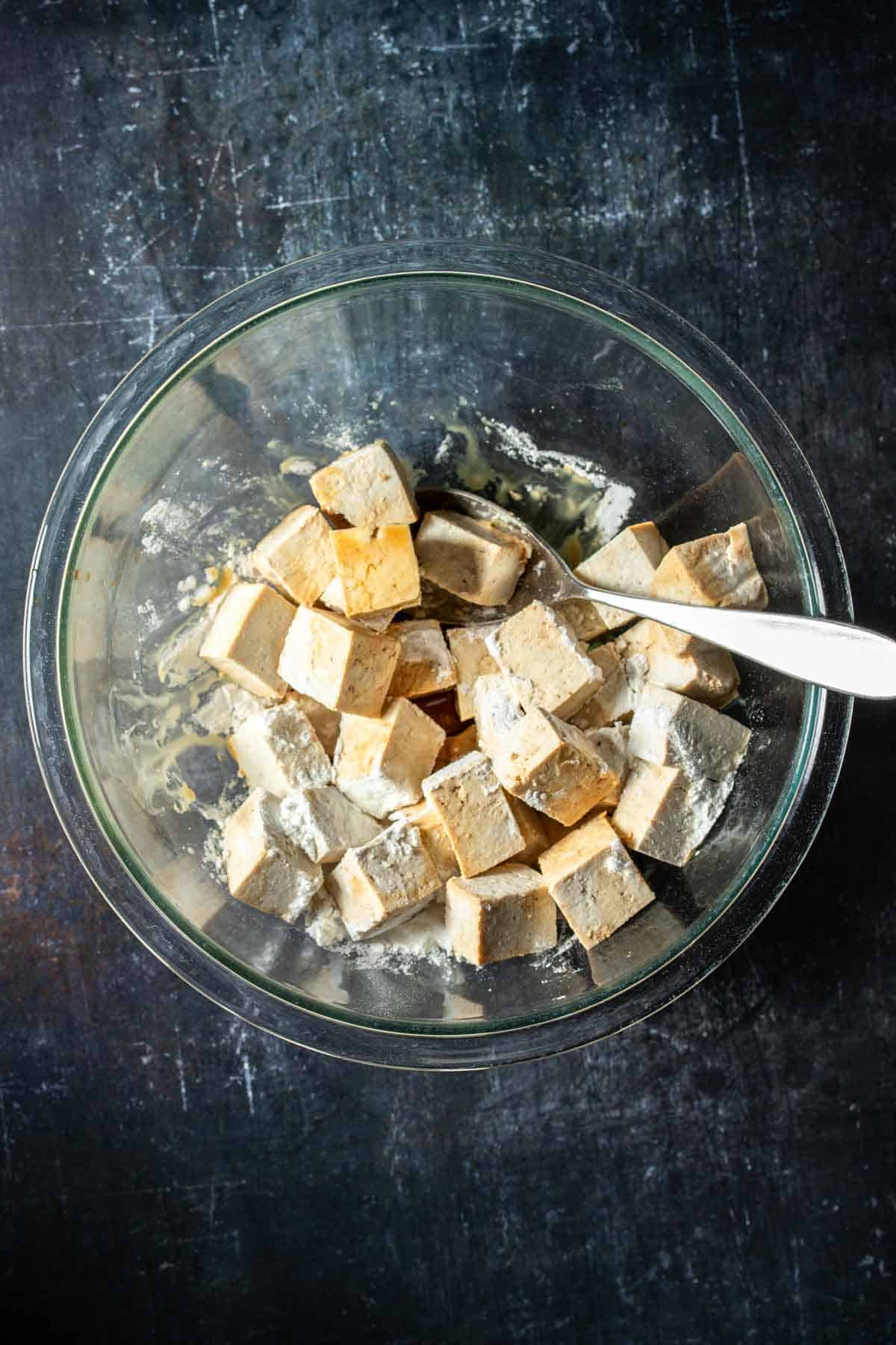 Spoon in a glass bowl of cut tofu pieces with cornstarch on them sitting on a dark surface.