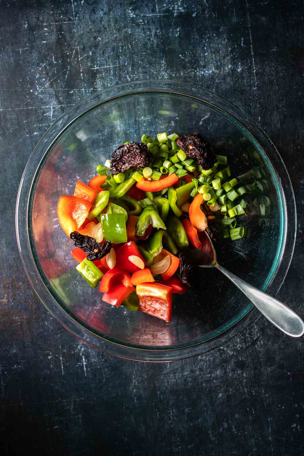 A glass bowl with cut green and red peppers and dried chilies inside and a spoon in it.