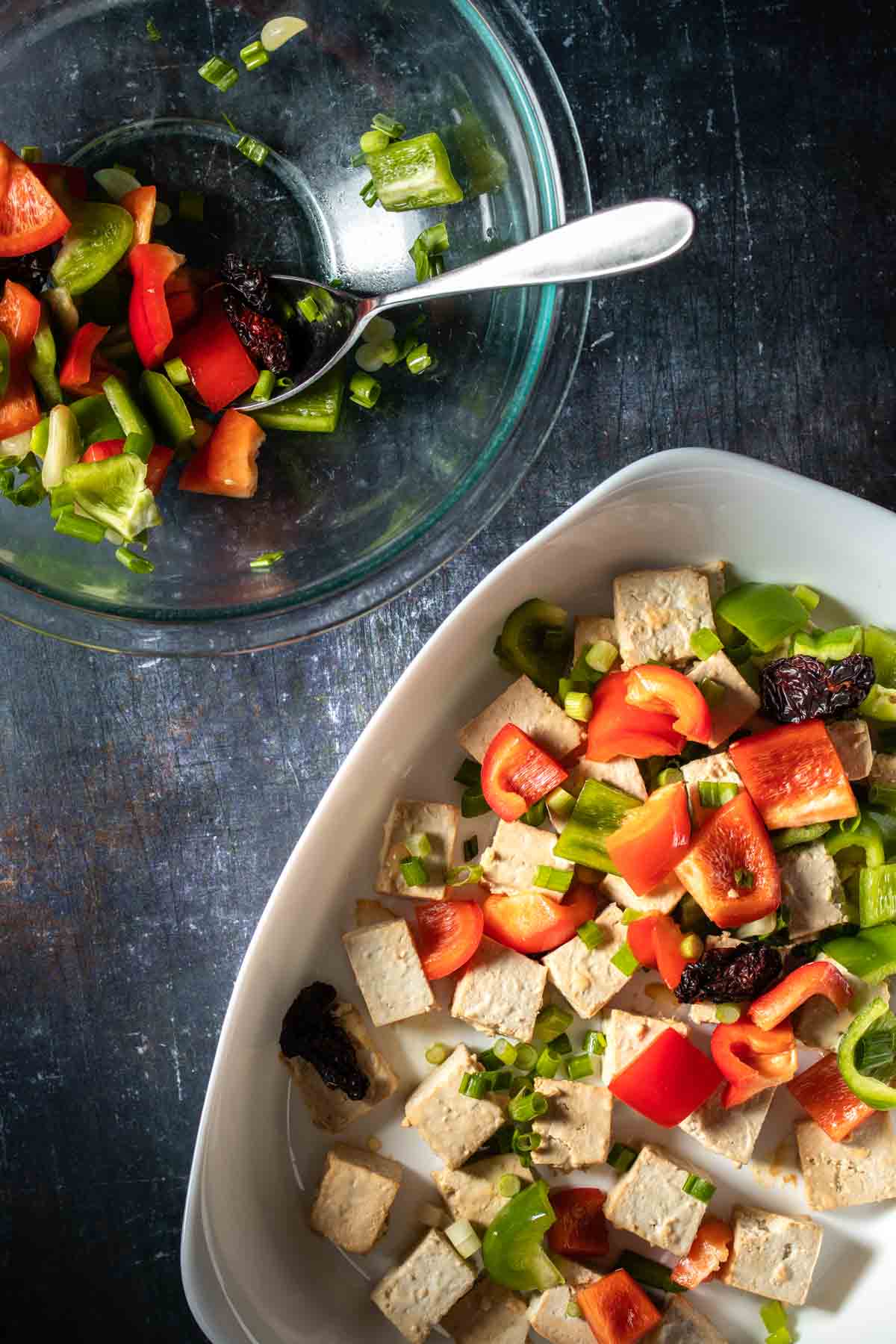 A white baking dish with squares of tofu topped with cut pieces of bell pepper next to a glass bowl of pepper pieces.