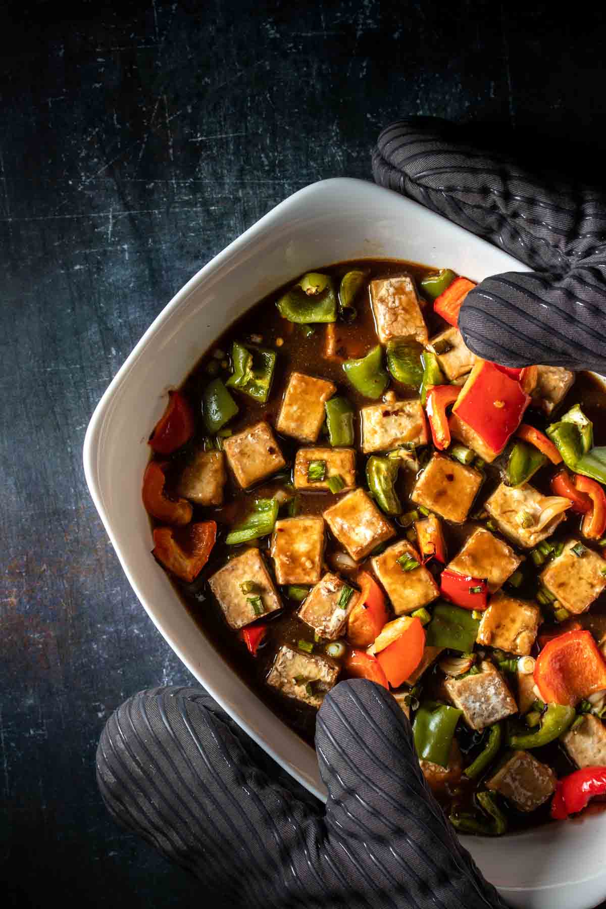 Grey oven mitts holding a white baking dish with baked tofu and red and green bell peppers and a brown sauce over them.