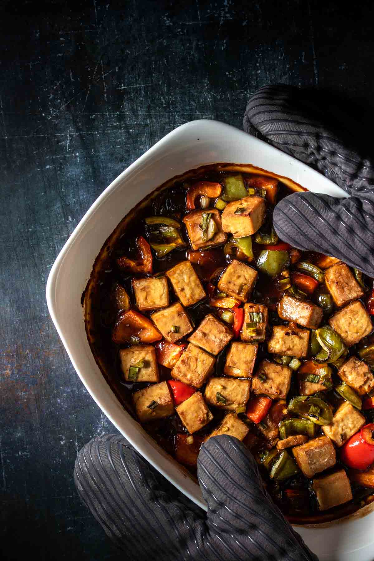 Cut pieces of tofu with red and green bell peppers and a thick brown sauce inside being held by grey oven mitts.