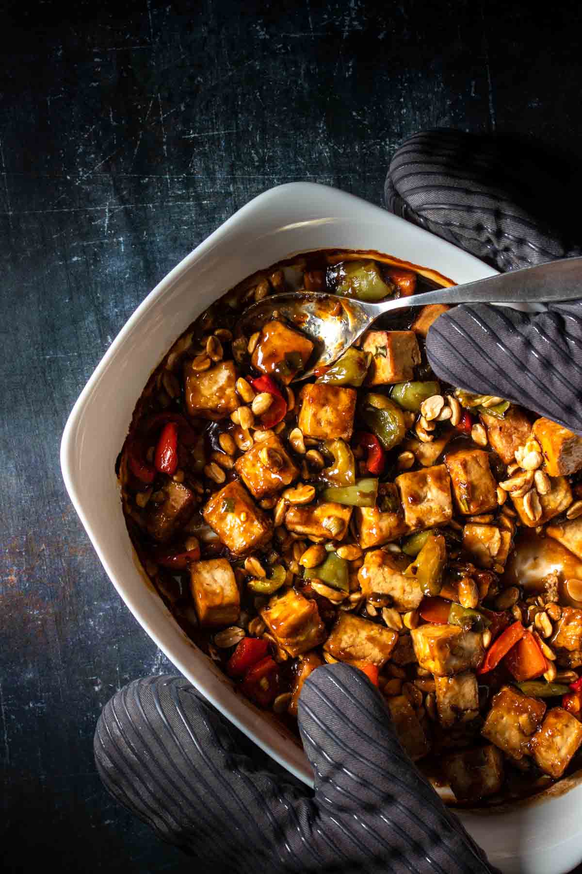 A Kung Pao tofu dish with red and green bell peppers mixed with peanuts baked in a white dish and held by grey oven mitts.