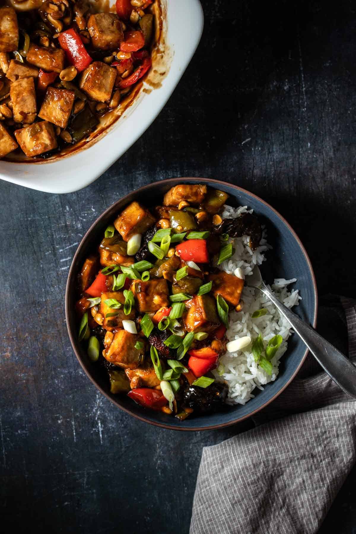 Top view of a dark blue bowl with white rice and Kung Pao tofu inside next to a white baking dish of it on a dark surface.