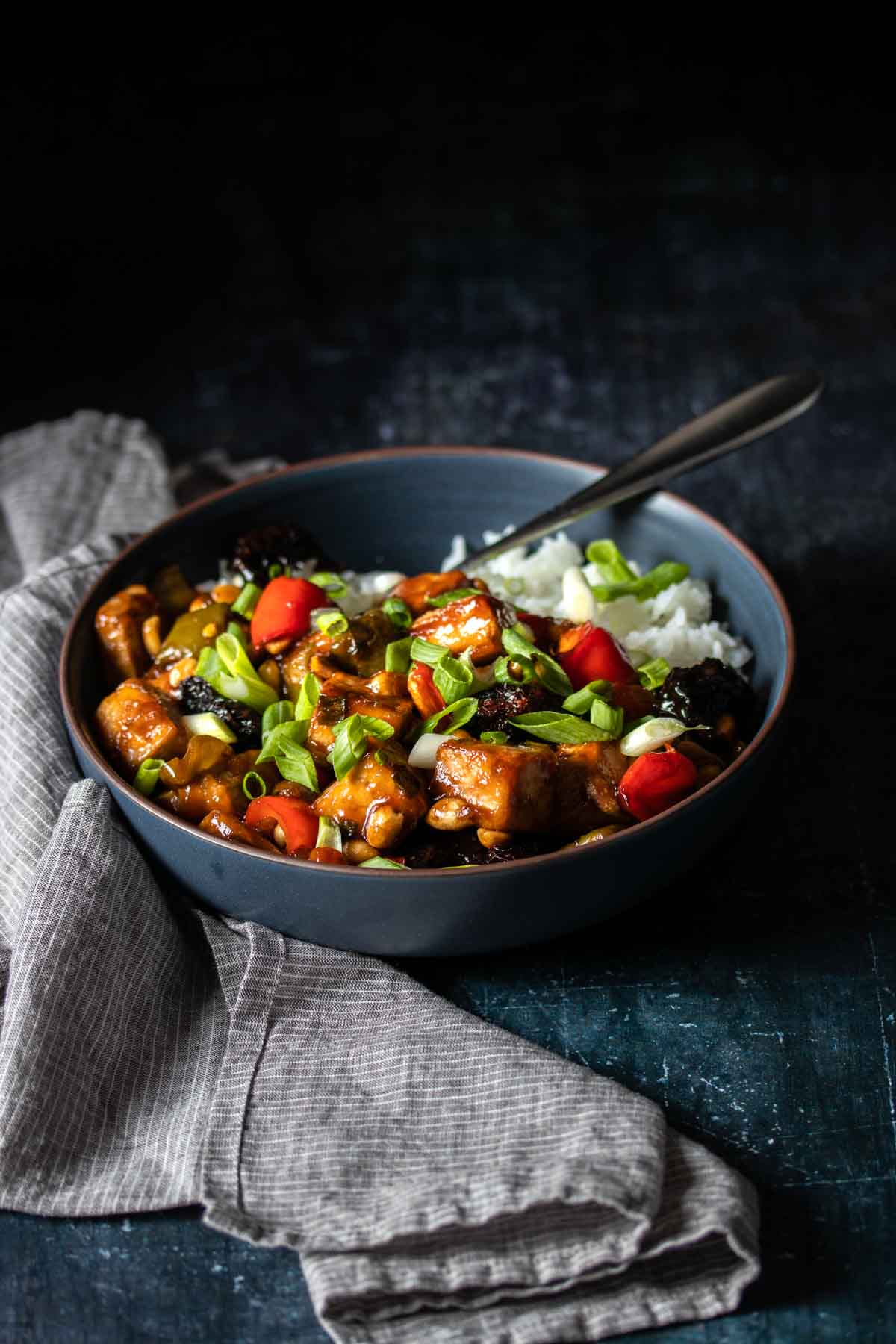 Front view of a dark blue bowl with white rice topped with Kung Pao tofu and sprinkled with green onions next to a grey towel.