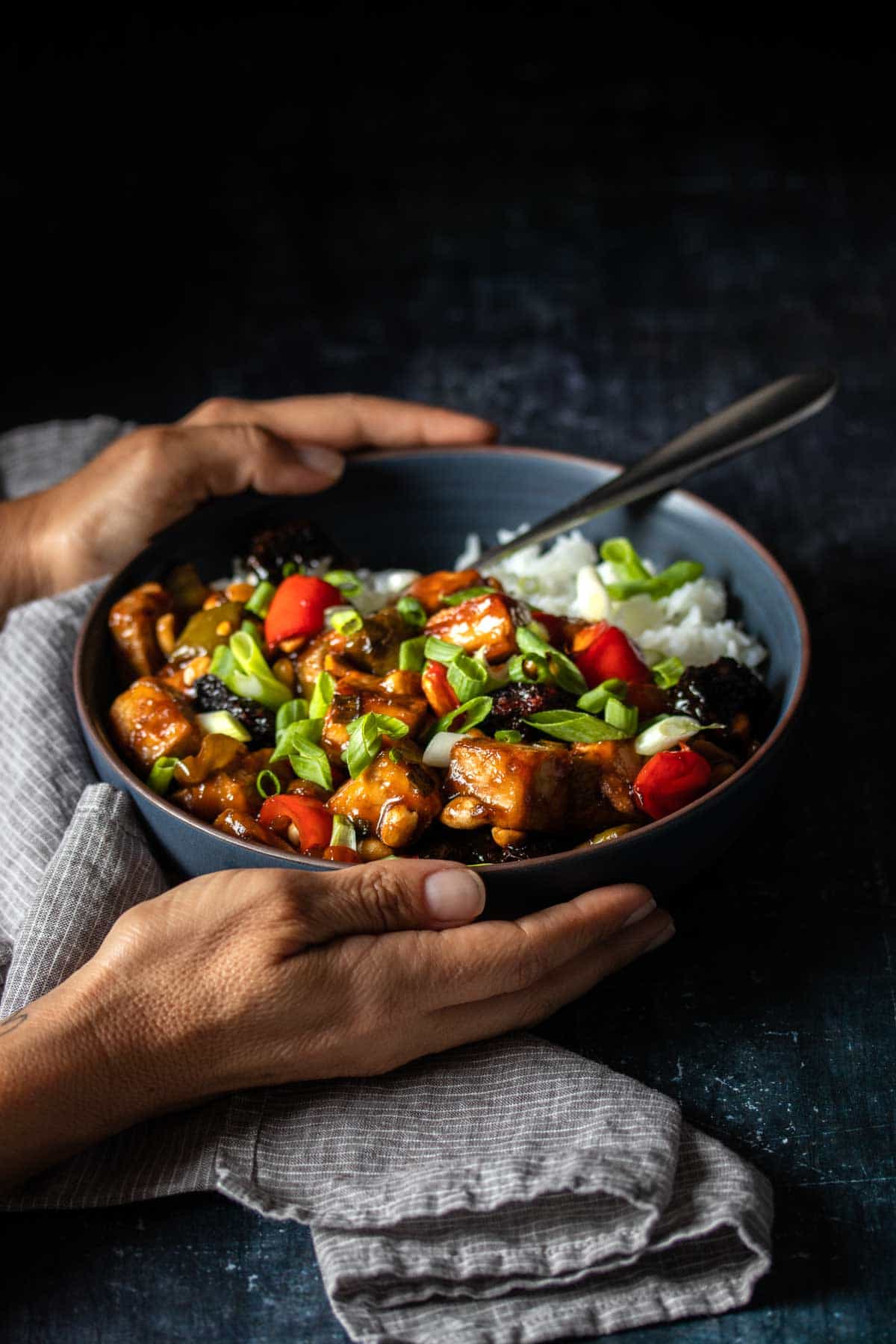 Two hands holding a dark bowl that has Kung Pao tofu over white rice inside and a grey towel under it.