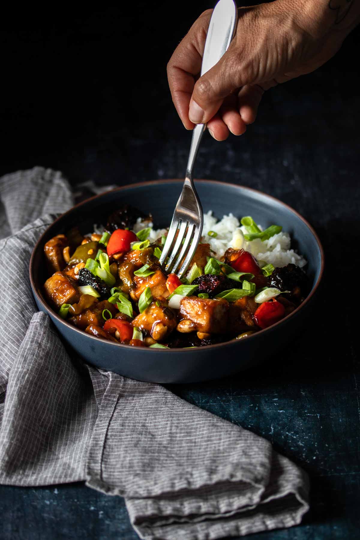 A fork getting a bite of Kung Pao tofu and white rice from a dark bowl next to a grey towel.