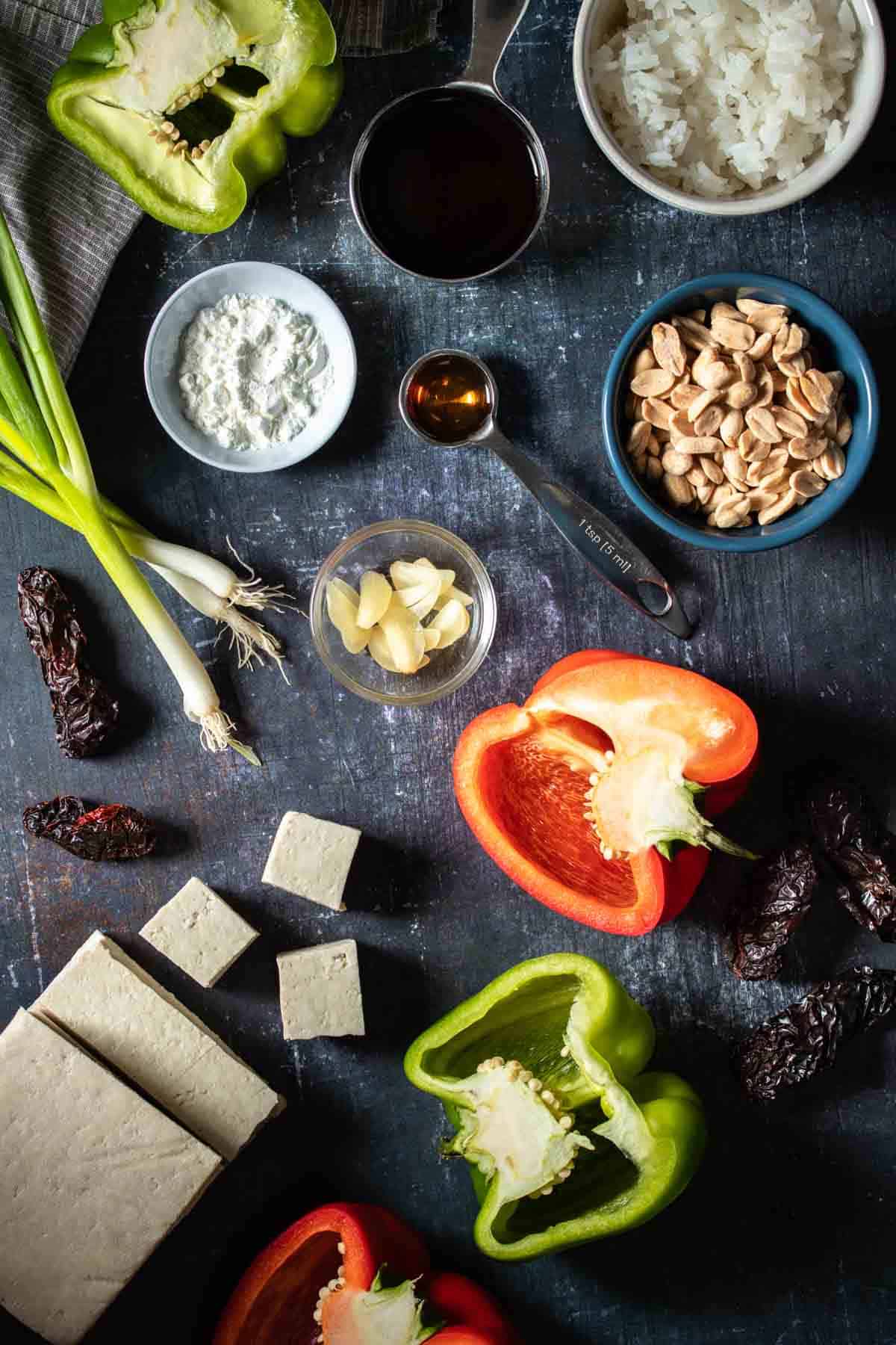 Top view of veggies, peanuts, cornstarch, tofu, and seasonings in measuring spoons and bowls.