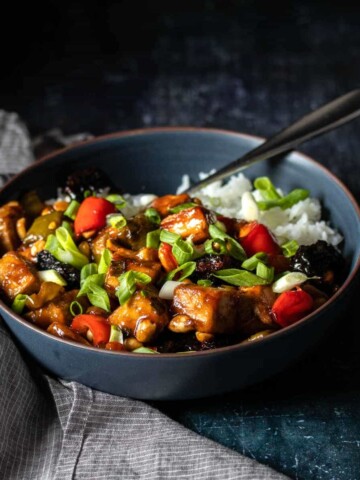 Kung Pao tofu over white rice in a dark blue bowl with a fork in it sitting next to a grey towel.