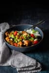 A grey towel twisted up next to a dark blue bowl with Kung Pao tofu inside over white rice.