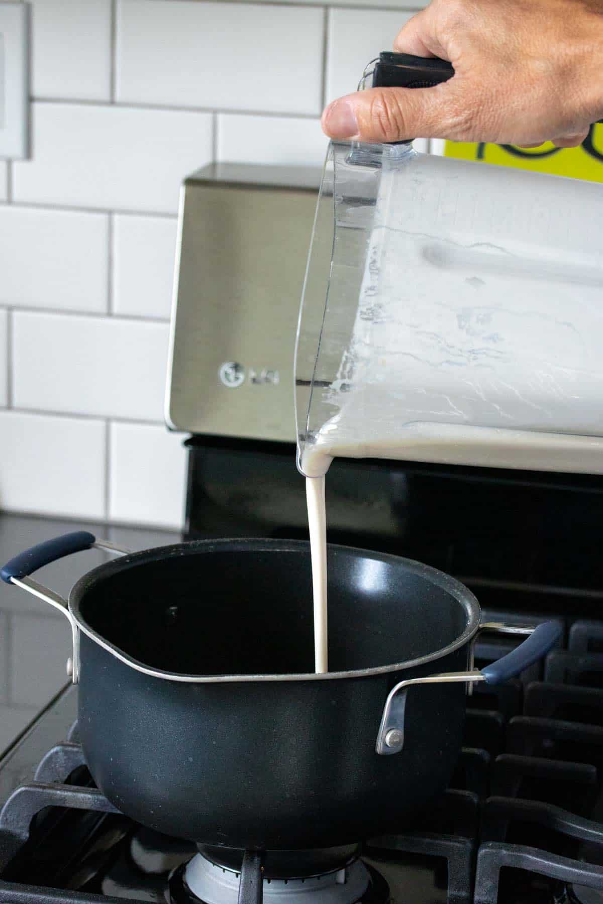 A hand pouring white liquid from a blender into a black pot on the stove.