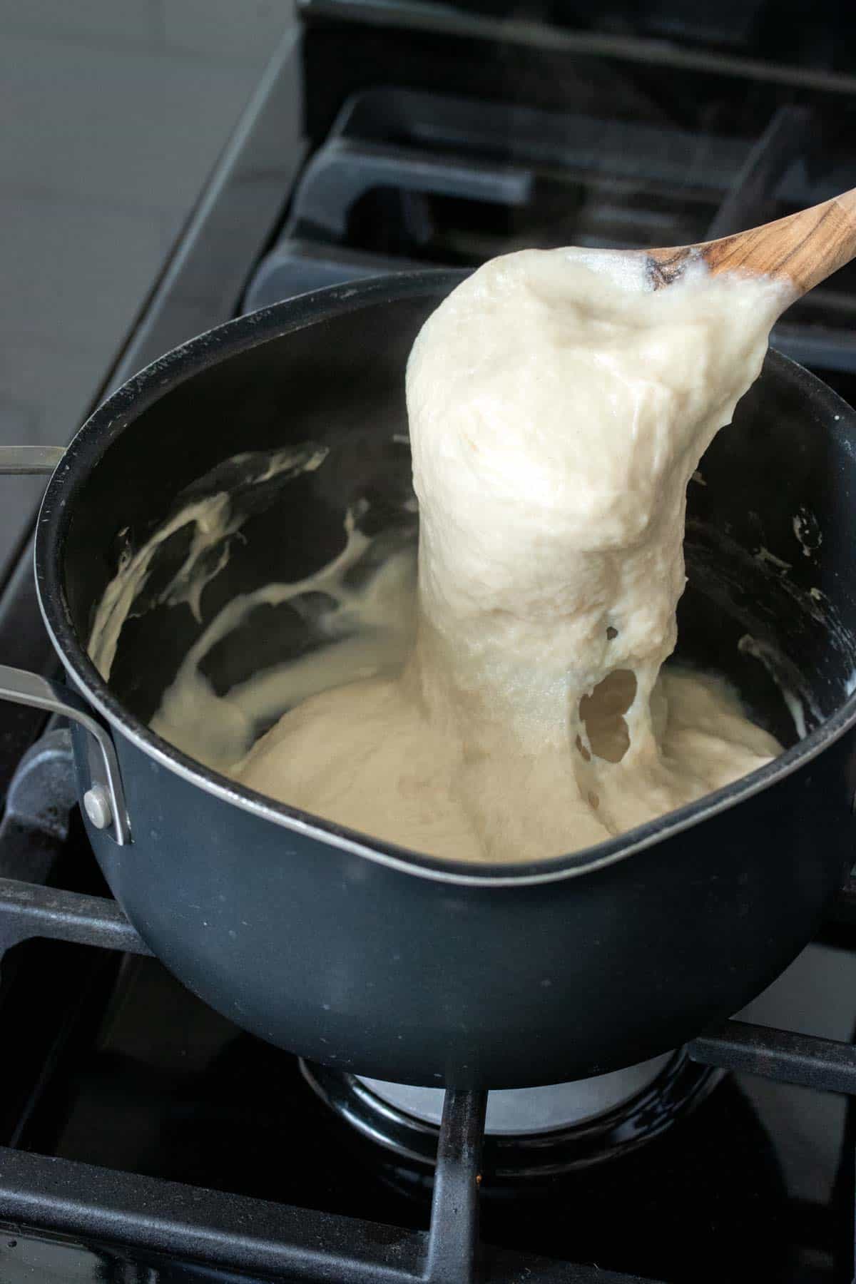 A wooden spoon mixing and picking up thick a gooey white cheese like substance from a pot.