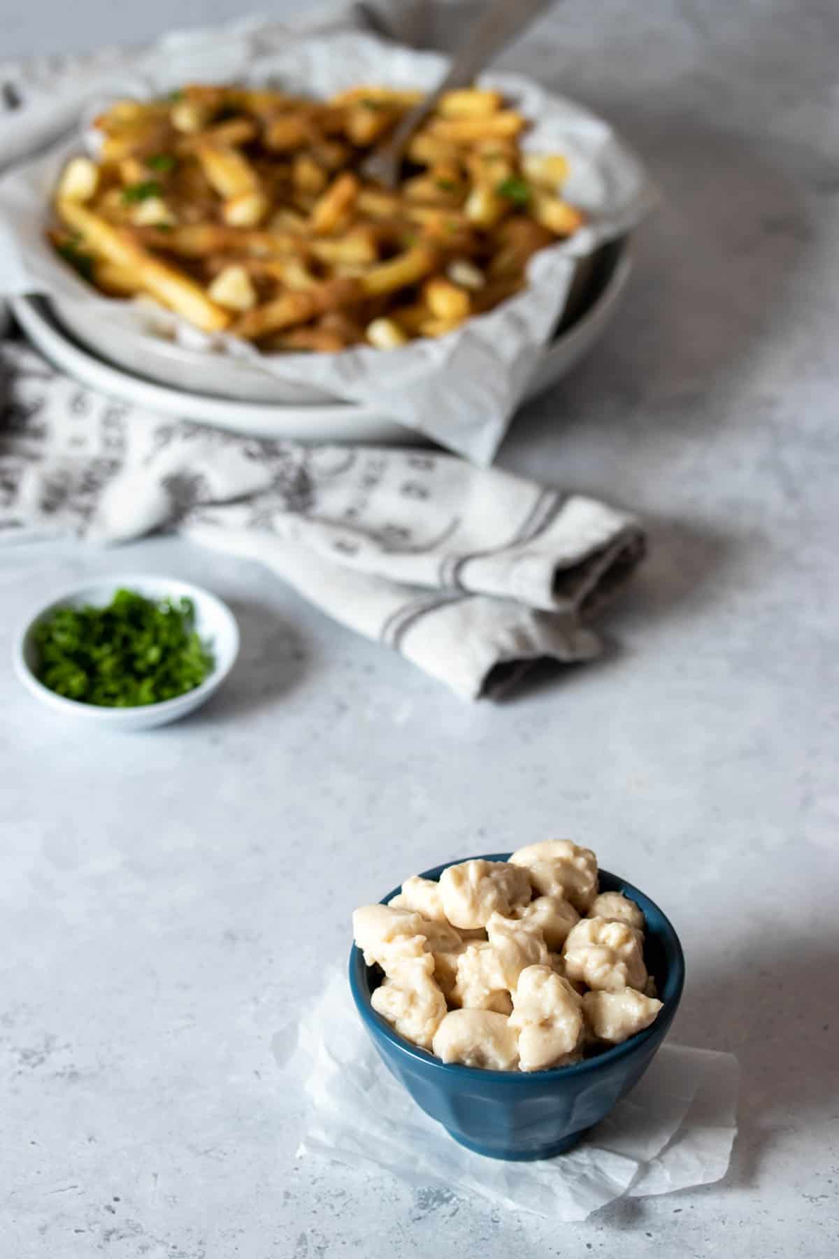 A small blue bowl with scoops of cheese curds in it and a bowl of poutine in the background.