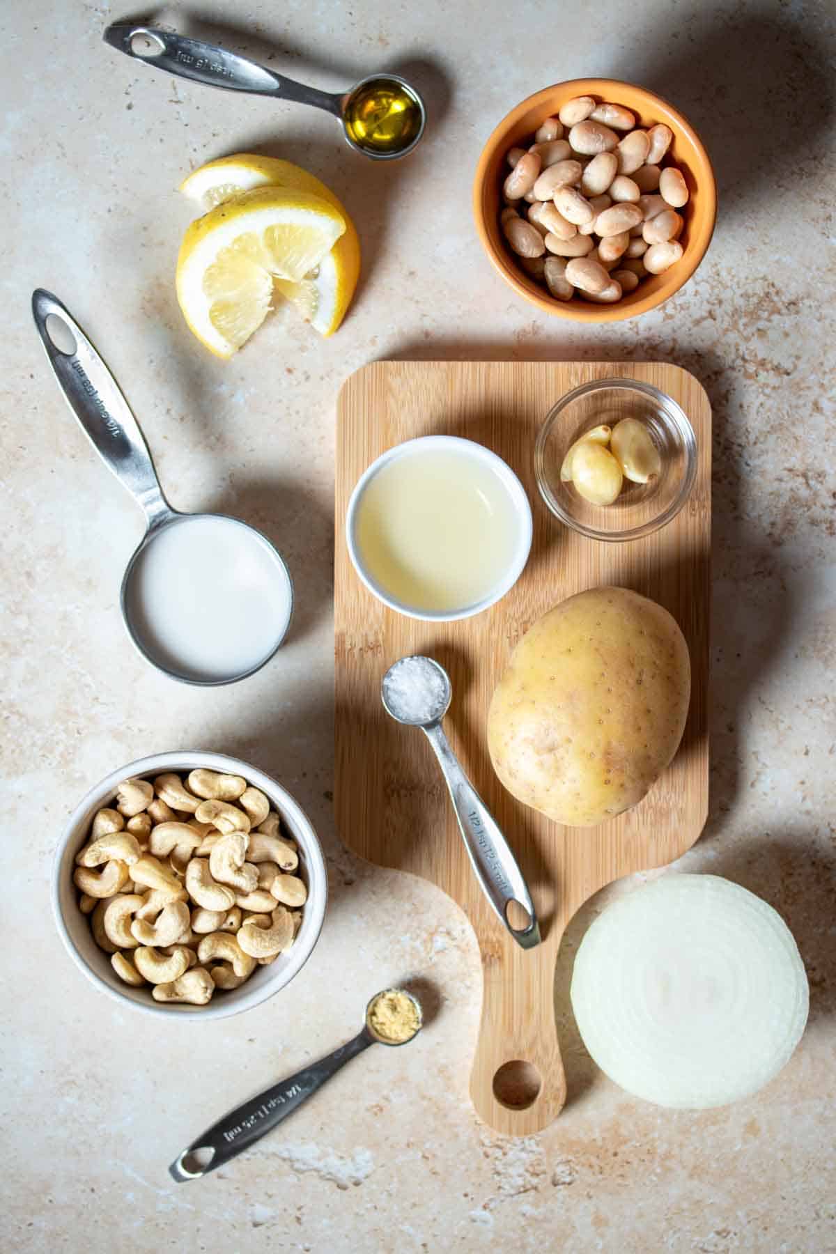 Top view of a tan surface with bowls of nuts, liquids, onoin, potato, white beans, lemon and seasonings.