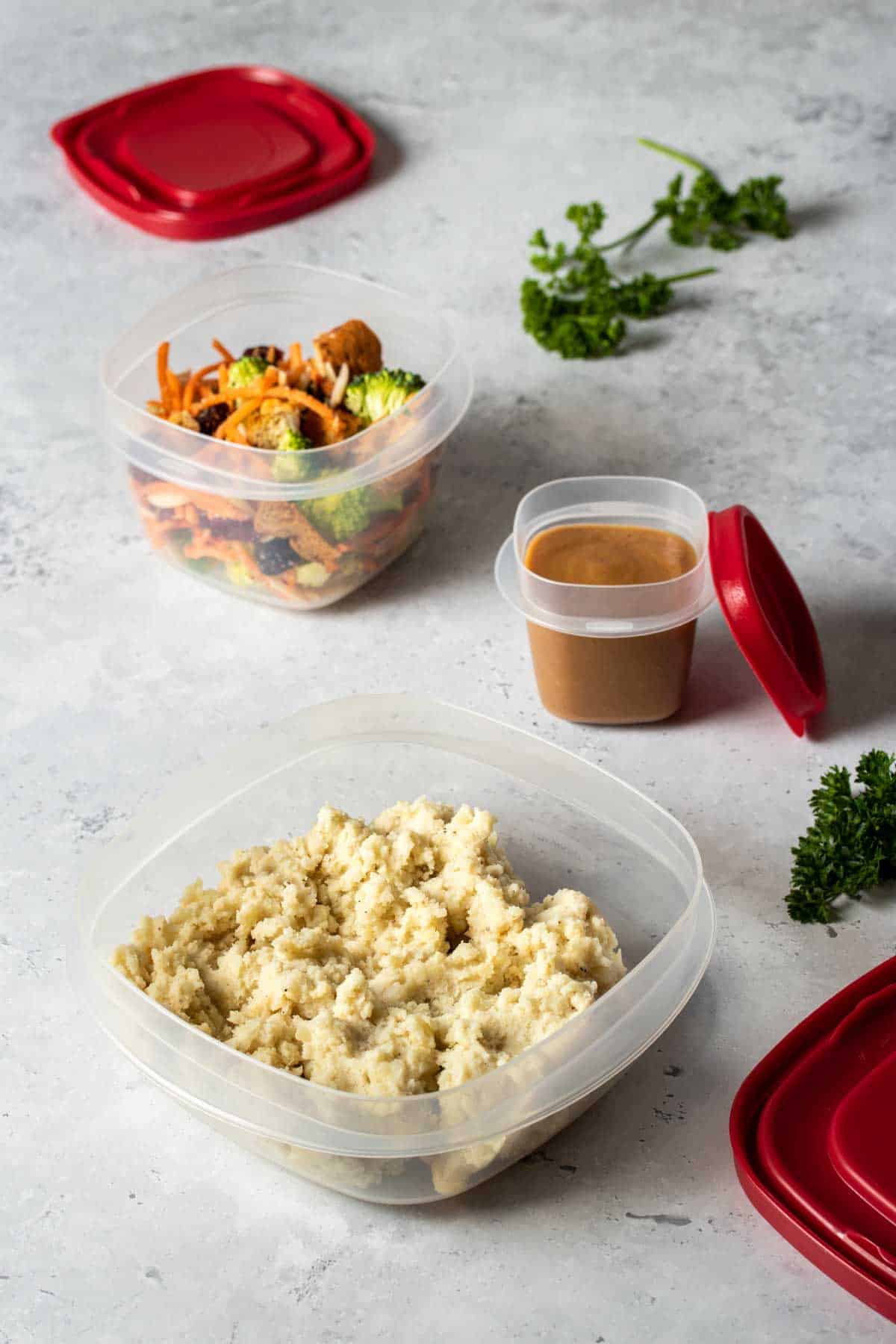 Three open plastic containers with red lids next to them filled with mashed potatoes, gravy and broccoli salad separately.