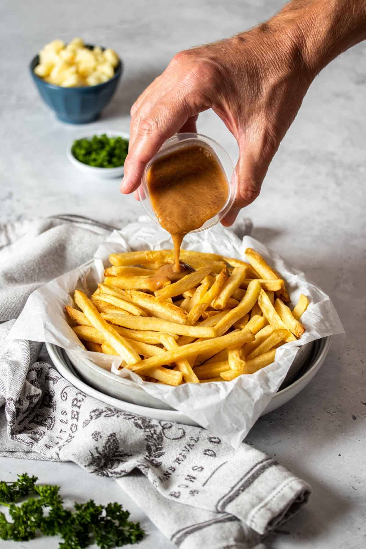 A hand pouring gravy from a plastic container onto a bowl of fries.