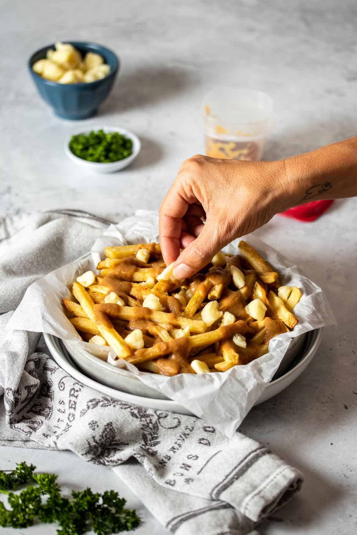 A hand putting cheese curds on a pile of french fries with gravy on them in a white bowl.