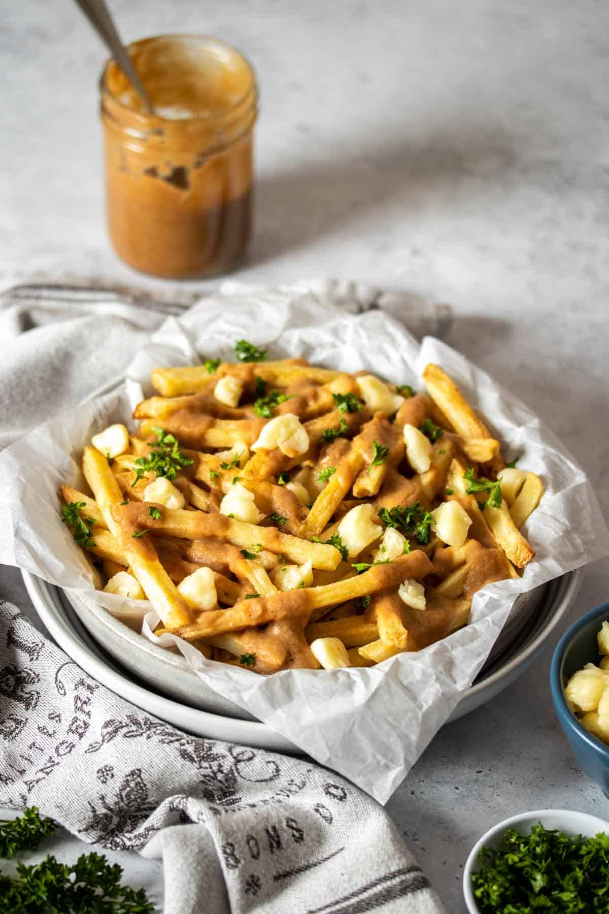 A stack of white bowls with parchment paper and poutine style french fries in them next to a towel and jar of gravy.