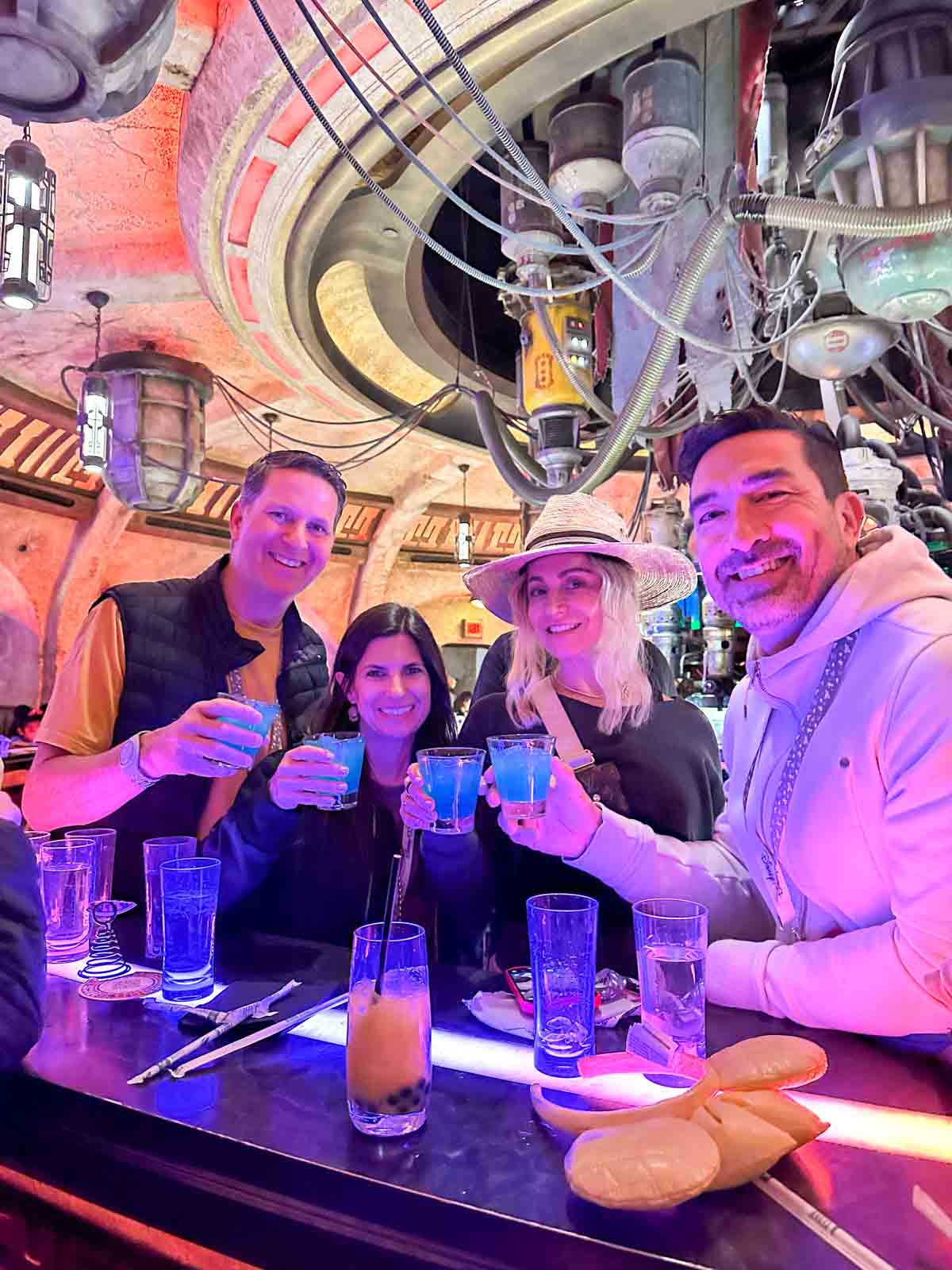 Two couples lit in pink purple light holding blue drinks over a bar top saying cheers.