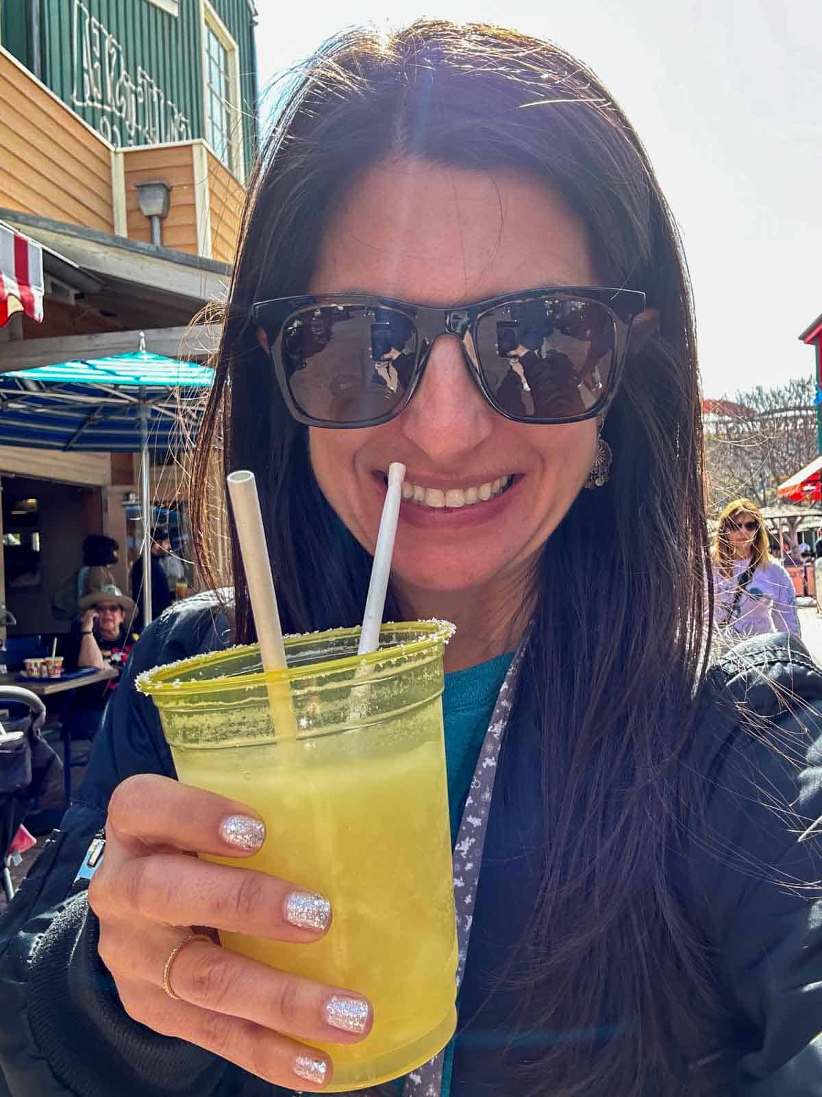 A woman with long brown hair holding a yellow plastic cup with two straws and a frozen margarita inside.