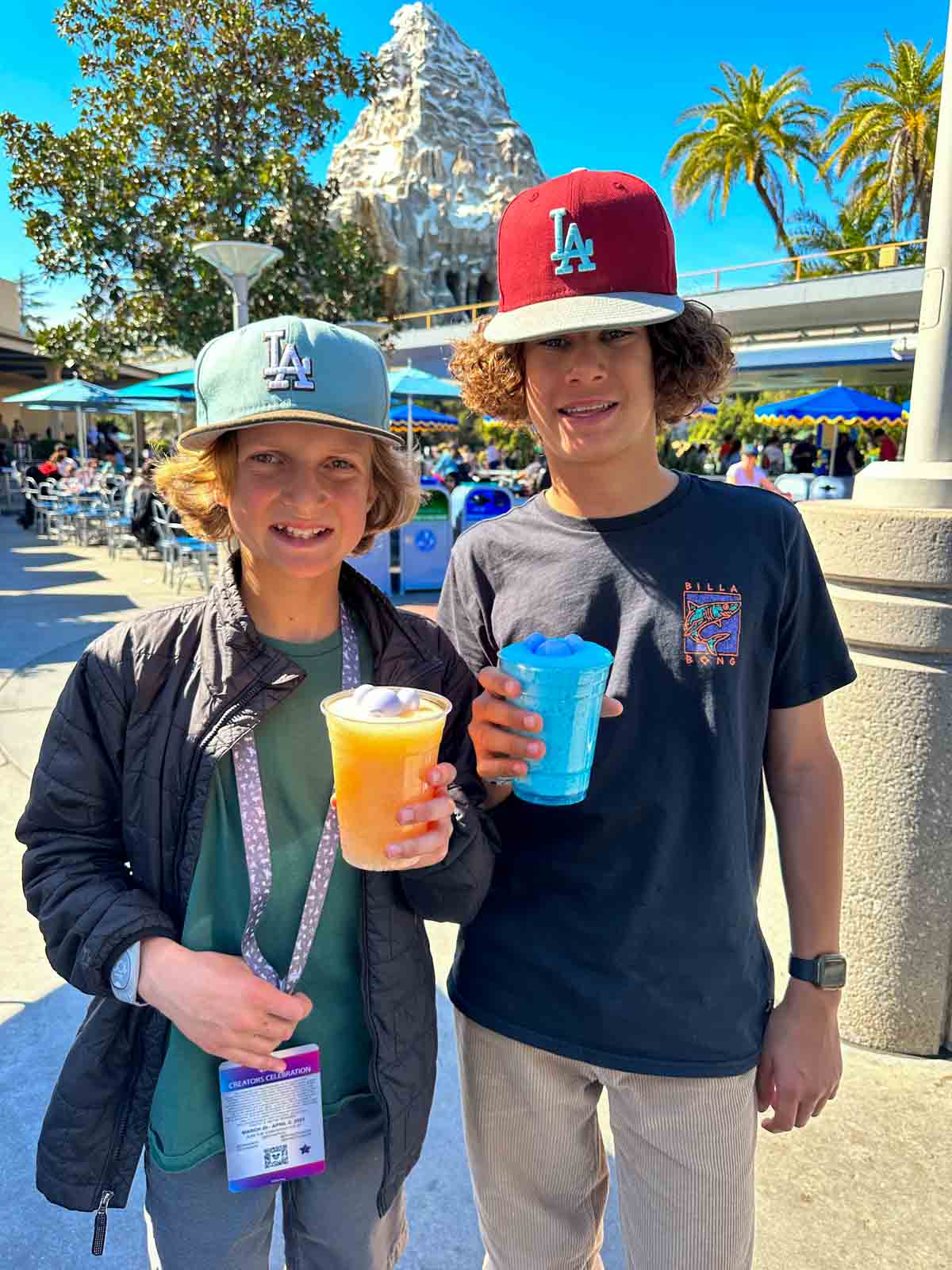 Two boys in hats holding yellow and blue frosties in plastic cups outside.