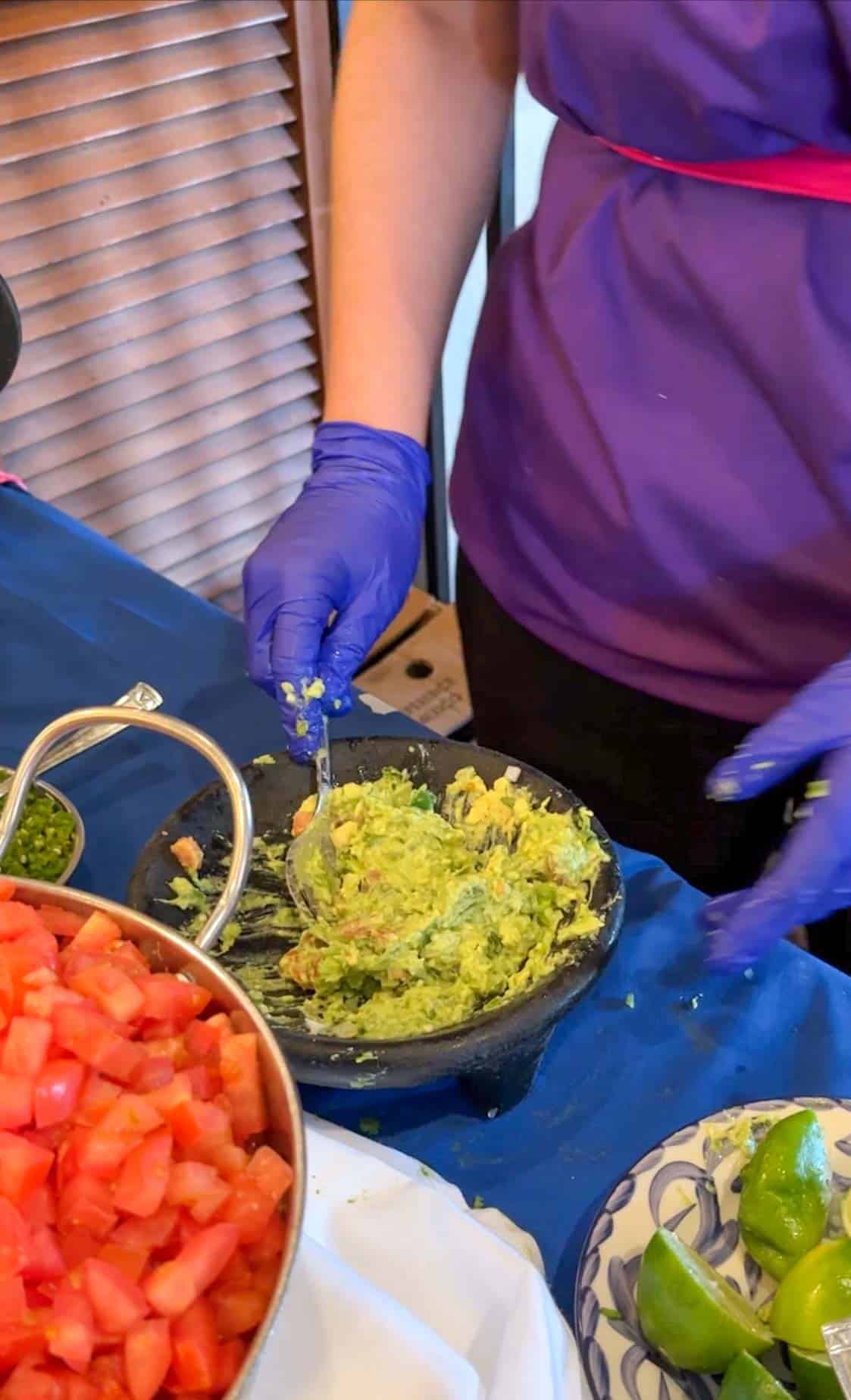 A hand with a purple plastic glove mixing guacamole in a black bowl on a blue table.